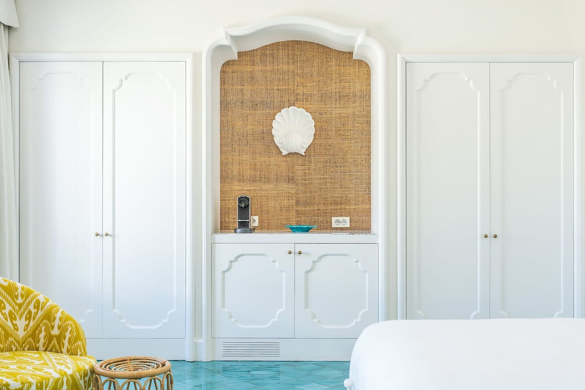 Una habitación con una cama, un sofá amarillo estampado, un sillón de mimbre, un armario blanco y una estantería de pared con un adorno y un cuenco azul.
