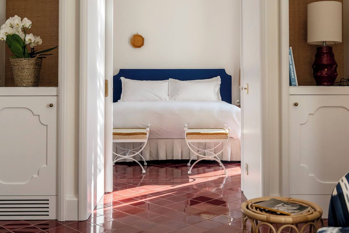 A bedroom with a bed, two bedside tables, a chandelier, and a wicker basket with flowers.