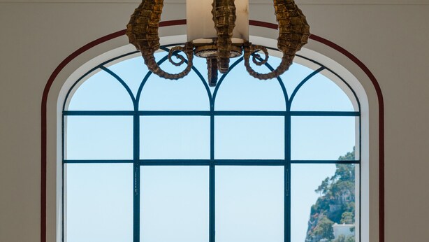 A room with a large arched window, a chandelier, a sofa, and a dining table. - Villa Helios Capri (Capri)