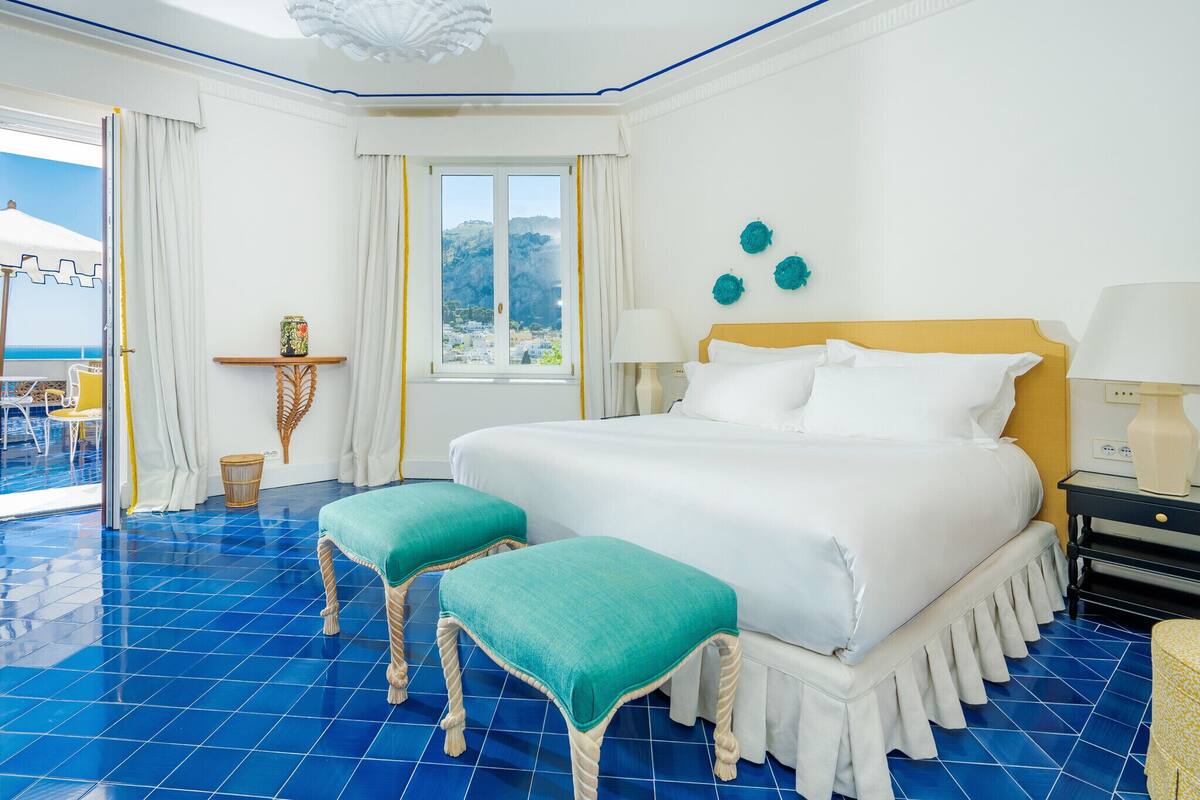 A hotel room with a large bed, two matching ottomans, a window with a view, a small table with a lamp, and a chair.
