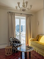 A room with a yellow sofa, a patterned armchair, a round table, a wicker stool, and a view through a large window.
