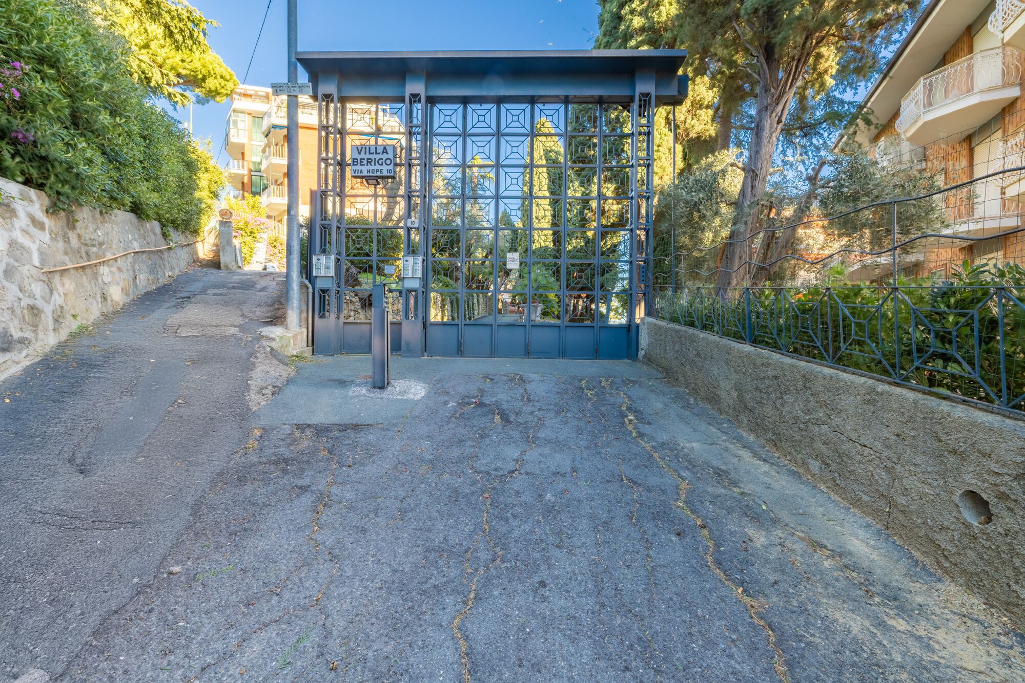 Ein begrenzter Zugang zur Villa Berigo, mit einem Metallgitter mit geometrischem Muster, einer Steinsäule links und einem von Bäumen gesäumten Weg.