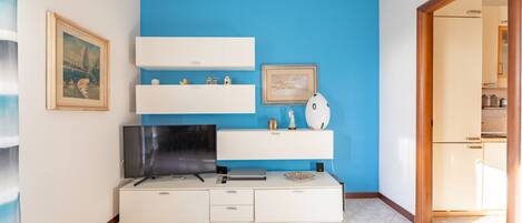 Una sala moderna con una pared azul de acento, un televisor de pantalla plana, un mueble blanco para equipo multimedia y una puerta que da a otra habitación.