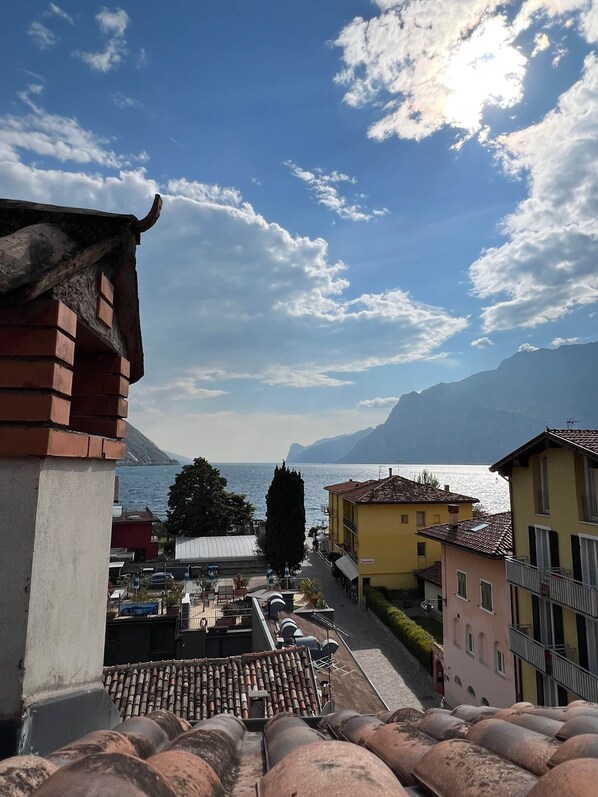 A view of a coastal town with terracotta-tiled rooftops, buildings with balconies, and a clear view of the sea and mountains in the background. - Attic 8 (Nago-Torbole)