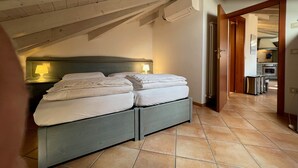 A bedroom with a bed, two bedside tables with lamps, a wooden ceiling, and a view into the kitchen. - Attic 8 (Nago-Torbole)