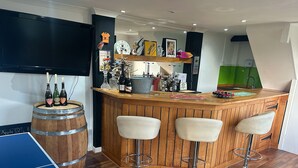 A bar area with wooden bar stools, a television mounted on the wall, and various bar items including bottles and glasses. - Luxury 5 Bedroom Group House near Cardiff with Sauna and Games Lounge (Penarth)