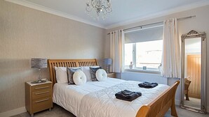 A bedroom with a wooden bed, a nightstand, a lamp, a mirror, and a window with curtains. - Luxury 5 Bedroom Group House near Cardiff with Sauna and Games Lounge (Penarth)