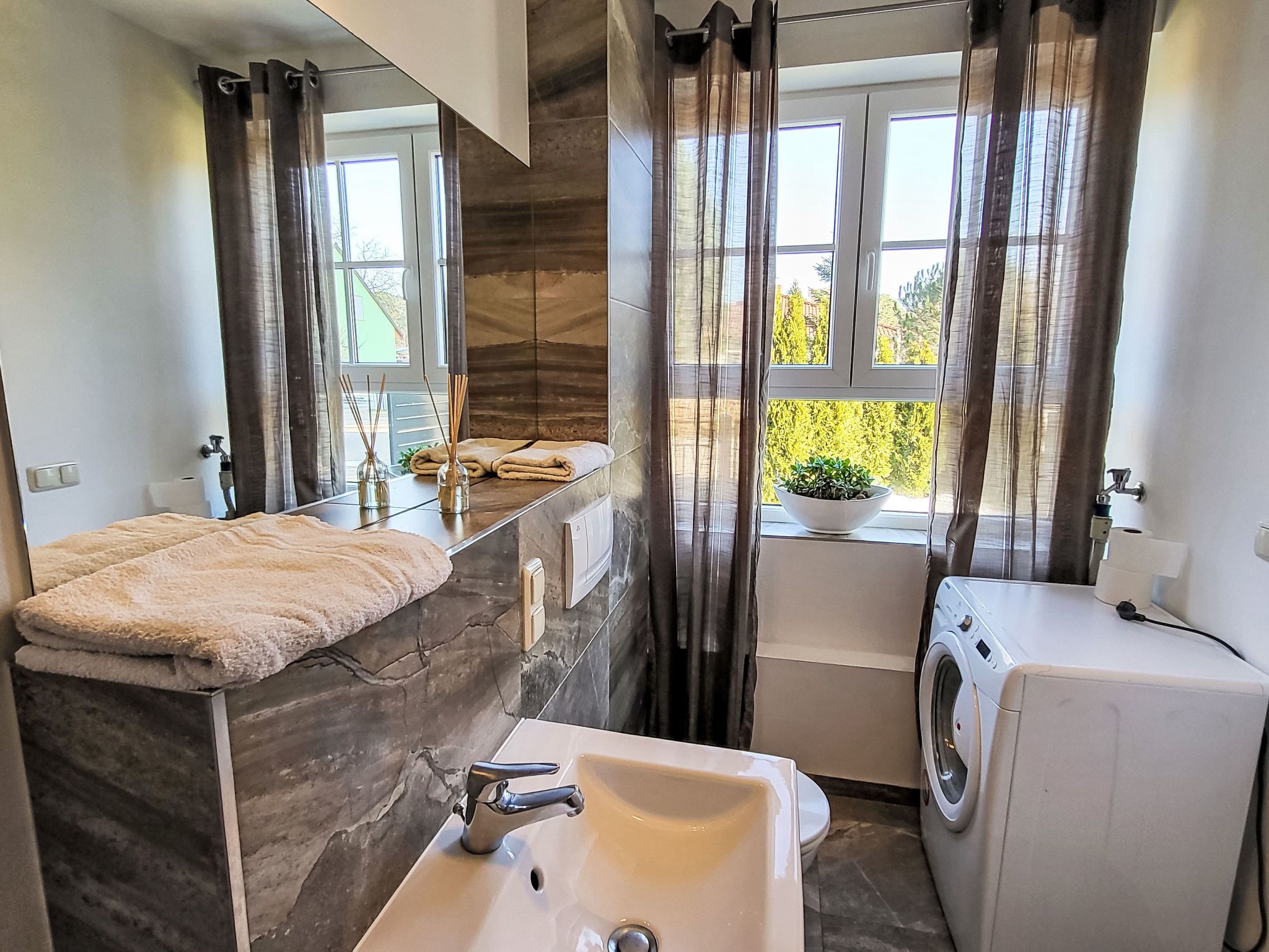 Une salle de bain moderne avec un comptoir en marbre, un lavabo, un lave-linge et une fenêtre donnant sur un paysage verdoyant.