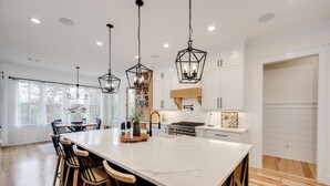 A spacious kitchen with a large island, wooden chairs, pendant lights, and white cabinetry.