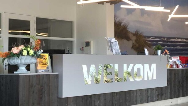 A reception desk with a 'WELCOME' sign, a potted plant, and a view of the ocean through a large window. - 2 minutes from the beach! Mobile home Duingras Ameland (Nes)