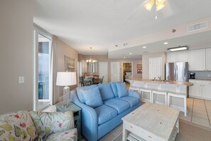 A modern living room with a blue sofa, a floral armchair, and a wooden coffee table. A kitchen with white cabinets and stainless steel appliances is visible in the background. - Lighthouse 1215 by Liquid Life (Gulf Shores)