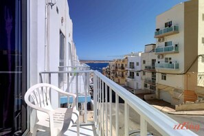 A balcony with a white plastic chair, a railing, and a view of a coastal town with buildings and the sea. - Sunny Holiday Apartment in Mellieha (Il-Mellieħa)