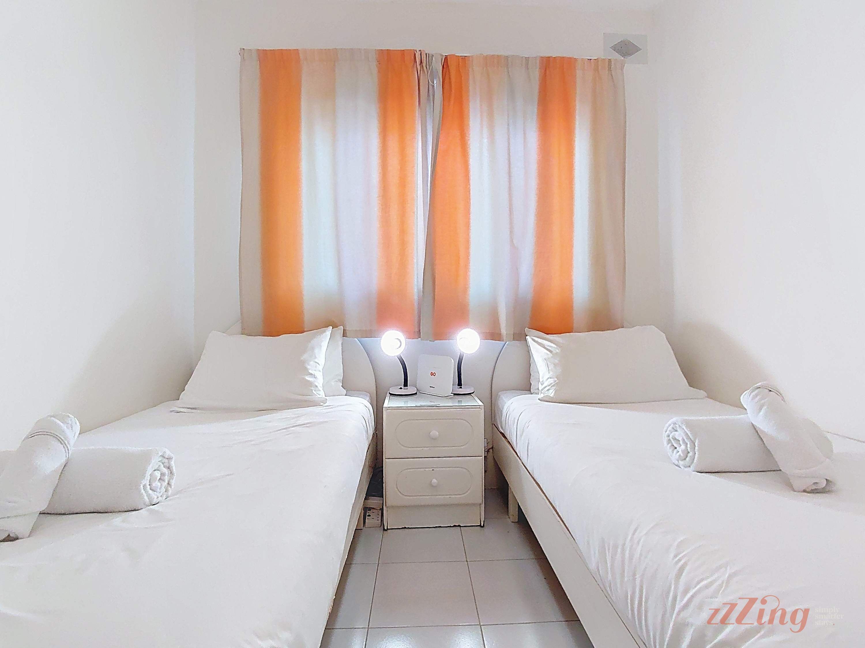 Une chambre d’hôtel avec deux lits simples, une table de chevet avec une lampe et un rideau à rayures orange et blanches.