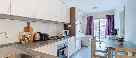 A modern kitchen with white cabinets, a stainless steel sink, a microwave, and a gas stove. There is a dining area with a table and chairs, and a window with purple curtains.