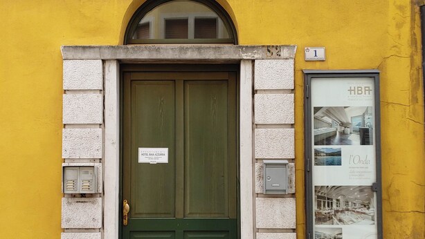 A green double door with a sign for Hotel Banca Azurra, a yellow wall, and a framed poster on the right. - Baia Terrazza 1 (Nago-Torbole)