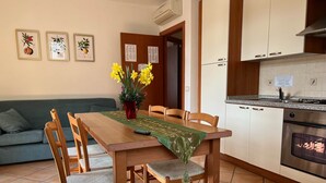 A dining area with a wooden table, chairs, and a vase of yellow flowers. There is a kitchen with white cabinets and a stove. - Baia Terrazza 1 (Nago-Torbole)