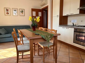 Un comedor con una mesa de madera, sillas y un florero con flores amarillas. Se observa una cocina con armarios blancos y una estufa.
