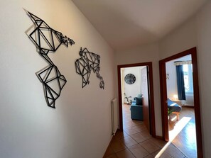 Un pasillo con arte mural geométrico, un reloj y vista a un salón con un sofá azul y una planta.