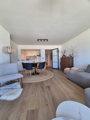 A modern living room with a dining area, a sofa, a coffee table, and a kitchen in the background. - Sea view apartment - Sorrente 4 (Blankenberge)