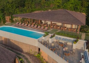 Un espace aménagé au bord de la piscine, avec des chaises longues, un coin salon abrité et un bâtiment doté d’un toit de chaume.