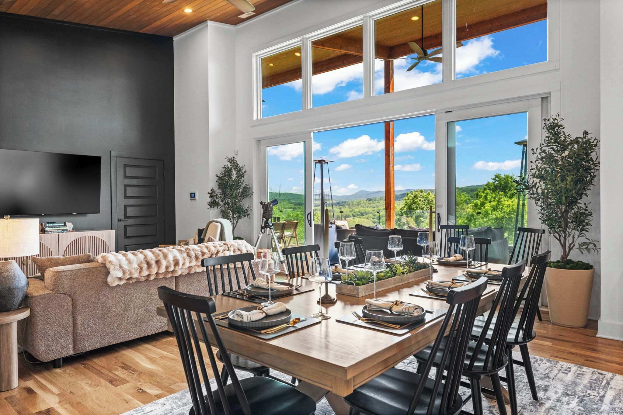 A modern dining room with a wooden table set for six, large windows offering a view of greenery and hills, a sofa with a fur throw, and a flat-screen TV on the left.
