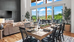 A modern dining room with a wooden table set for six, large windows offering a view of greenery and hills, a sofa with a fur throw, and a flat-screen TV on the left.