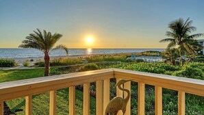 Una terraza de madera con barandilla, con vista a una playa con palmeras y un atardecer.