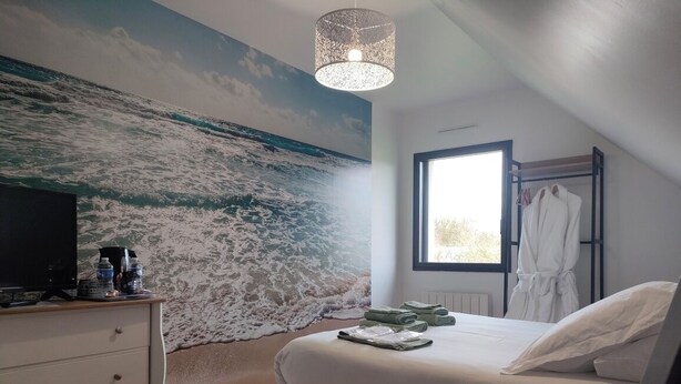 A bedroom with a large wall mural of the ocean, a bed with white linens, a bedside table with a lamp, and a window with a view of the outdoors.