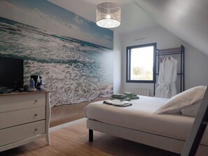 Et soveværelse med et stort vægmaleri af havet, en seng med hvide sengetøj, et natbord med en lampe og et vindue med udsigt til det fri.