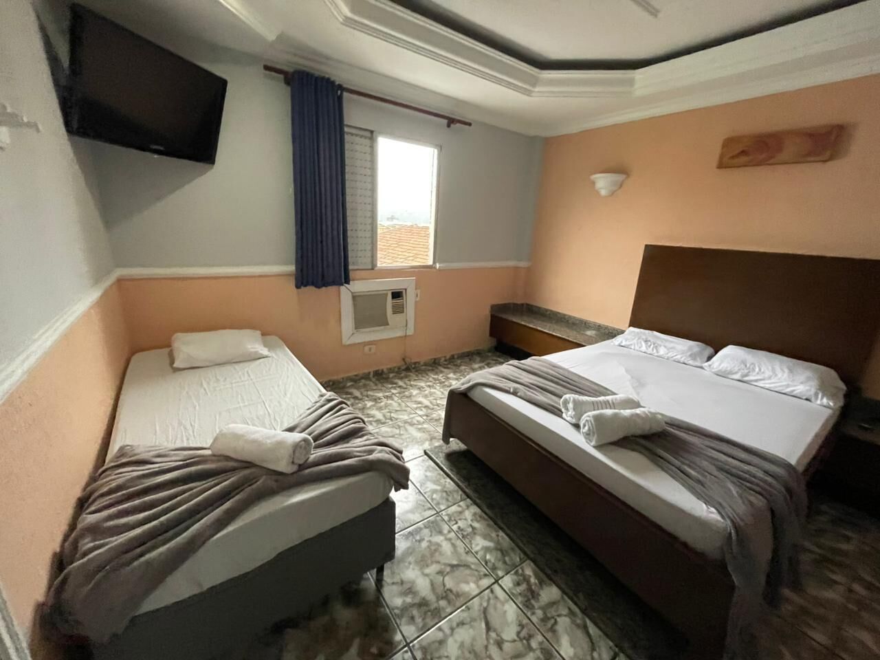 Habitación de hotel con dos camas, televisor, ventana con cortinas y aire acondicionado instalado en la pared.