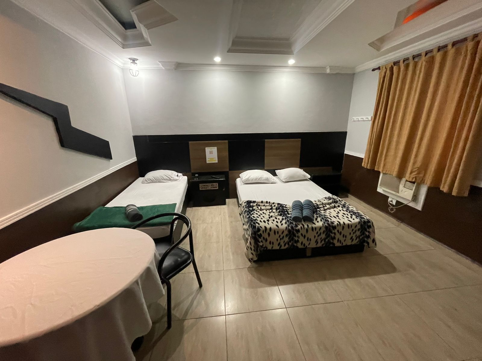 Habitación de hotel con dos camas individuales, una mesita pequeña, una silla, un reposapiés estampado y una lámpara en el techo.