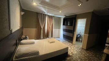 Una habitación de hotel con cama, cortinas, televisor y un baño visible a través de una puerta abierta.