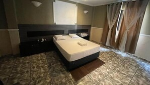 Une chambre spacieuse avec un grand lit, une tête de lit, un téléviseur à écran plat et une fenêtre avec des rideaux.
