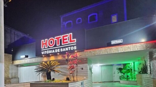 Un hotel llamado "HOTEL VITORIA DE SANTOS" con una fachada moderna, un cartel que indica la entrada y un área de recepción bien iluminada.