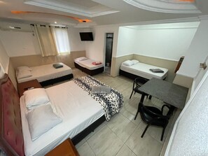 Ein Hotelzimmer mit drei Betten, einem Schreibtisch, einem Stuhl, einem Fernseher und einem Vorhangfenster.