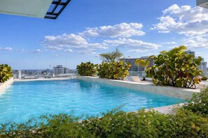 Ein Pool auf dem Dach mit Blick auf die Stadt, umgeben von Grünpflanzen und Palmen.