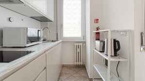 A compact kitchen with a microwave, toaster, and kettle on shelves, a window with a patterned curtain, and a white radiator. - GuestHost - A comfortable 30 square meters studio, perfect for two people, located on the first floor of a building with an elevator (NOT suitable for people with reduced mobility).The apartment is just steps from Pasteur metro station and within a 10-mi (Milan)