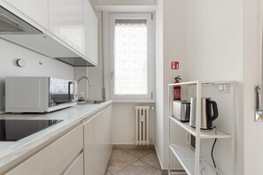 Angolo cottura compatto con forno a microonde, tostapane e bollitore su mensole, una finestra con tenda decorata e un termosifone bianco.