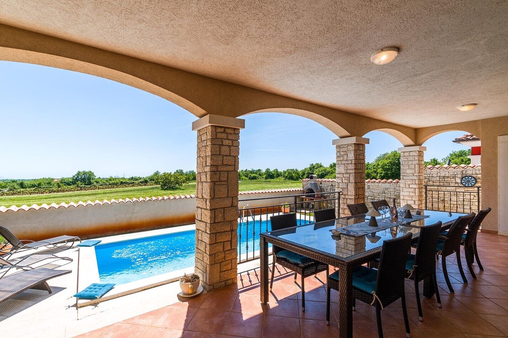 Un patio exterior con piscina, mesa de comedor y sillas, rodeado de pilares de piedra y vistas al paisaje.