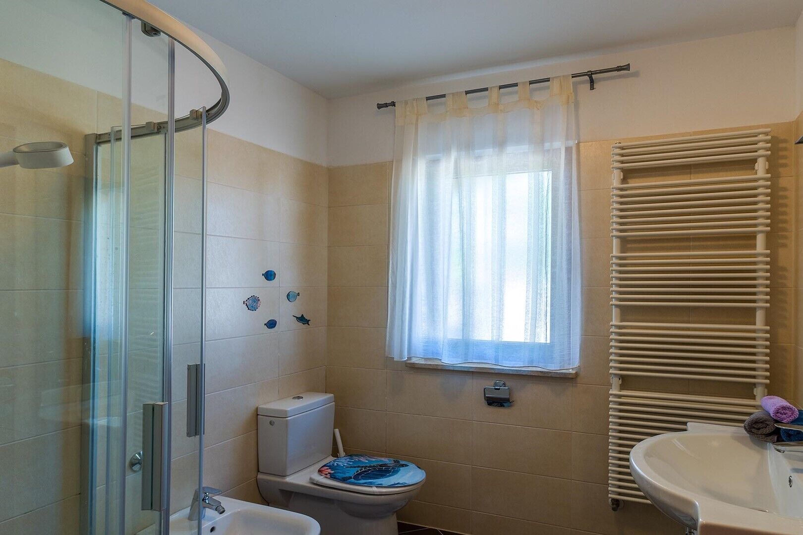 A bathroom with a glass shower enclosure, a toilet, a window with sheer curtains, a towel rack, and a sink.