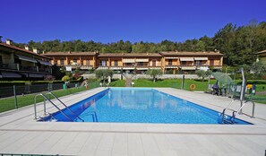 A swimming pool with a clear blue water, surrounded by a tiled deck. In the background, there are multi-story buildings with balconies and a green lawn. - Appartamento Borgo Alto (Puegnago del Garda)