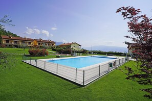 A rectangular swimming pool surrounded by a metal fence, with a well-maintained lawn and a large building in the background. - Appartamento Borgo Alto (Puegnago del Garda)