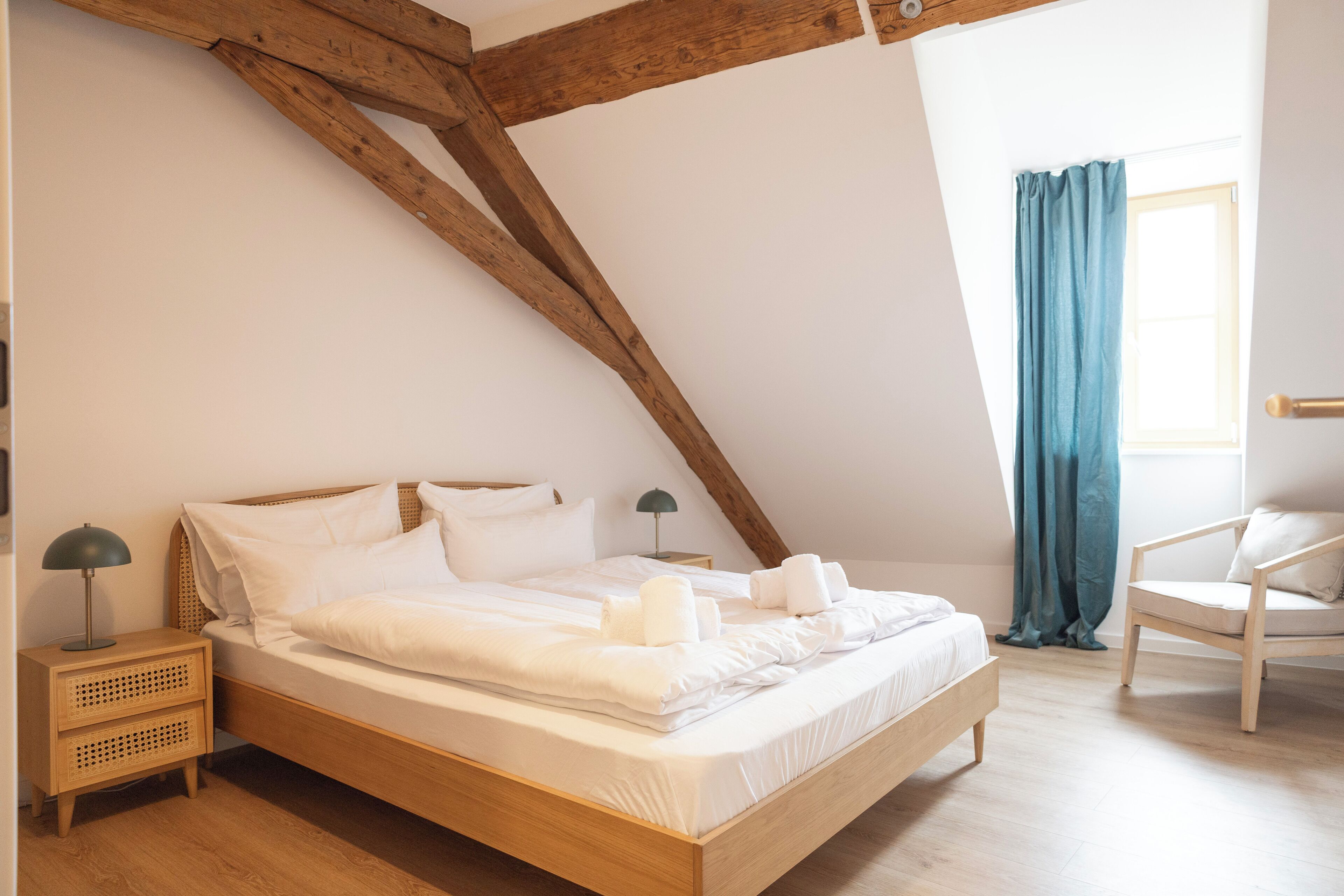 A bedroom with a wooden ceiling, a bed with white linens, a nightstand with a lamp, and a chair.