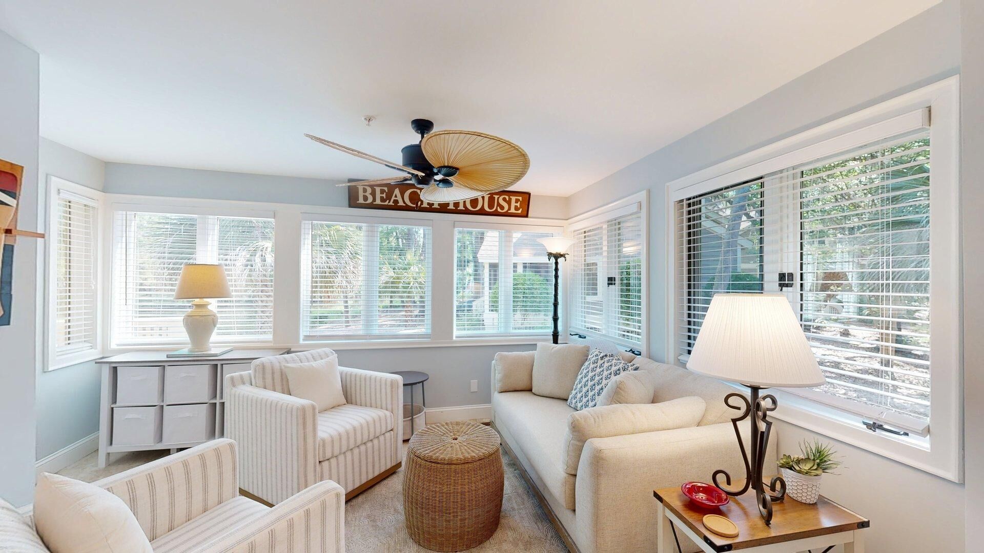 A living room with a ceiling fan, a sofa, a chair, a lamp, and a sign that reads 'BEACH HOUSE'.
