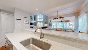 Una cocina con un lavamanos de acero inoxidable, un comedor con candelabro y una sala de estar con un ventanal grande.
