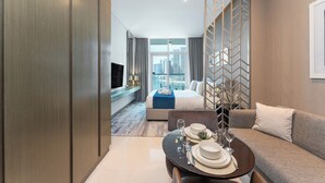Ett modernt hotellrum med en säng, en soffa, ett matbord med tallrikar och glas, och utsikt över stadsbilden genom stora fönster.
