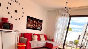 Una sala de estar con un sofá rojo, una mesa de centro blanca, un microondas rojo y un refrigerador pequeño. Hay un reloj en la pared y un cuadro sobre el sofá.
