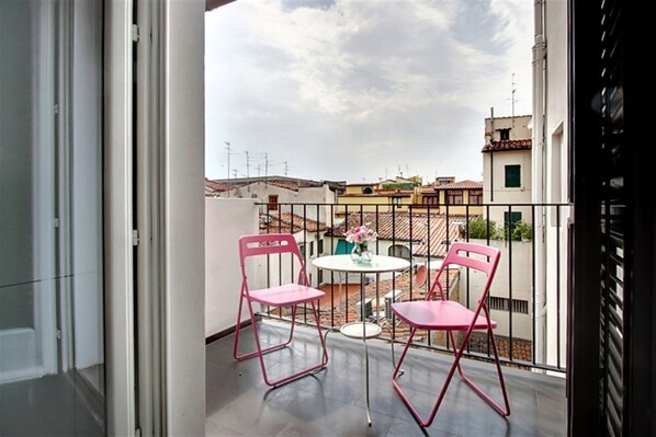 Un balcon avec deux chaises roses et une petite table, donnant sur un paysage urbain avec des immeubles et un ciel nuageux.