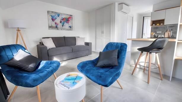 Un salon moderne avec un canapé gris, deux fauteuils bleus, une table basse blanche avec des magazines, un lampadaire et un coin cuisine doté d’un comptoir-bar.