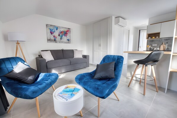 A modern living room with a grey sofa, two blue armchairs, a white coffee table with magazines, a floor lamp, and a kitchen area with a bar counter.